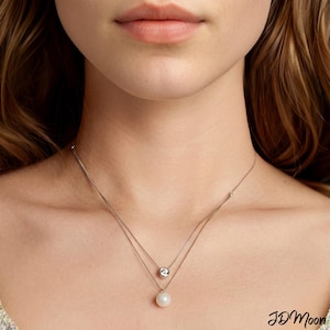 Puede incluir: Un delicado collar de plata con dos hilos. El hilo superior presenta una pequeña gema transparente, mientras que el hilo inferior tiene un colgante de perla blanca. El collar se muestra sobre un fondo de tono de piel.