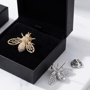 Peut inclure: Broche abeille dorée ornée de cristaux, présentée dans un écrin noir. Une broche abeille argentée et une épingle sont également visibles. Les broches présentent des ailes détaillées, parfaites pour accessoiriser.