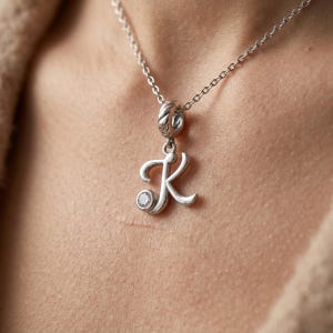 Könnte beinhalten: Eine silberfarbene Halskette mit einem Anhänger in Form eines kursiven Buchstaben "K". Der Anhänger hat einen kleinen, klaren Edelstein. Die Kette ist filigran und der Anhänger hängt an einem dekorativen Bügel.