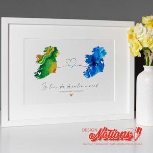 May include: White framed art print with abstract watercolor maps of Ireland in green and blue, linked by a heart. The print includes the text "Is leor don deoclin a nend" and "HOME IS WHERE THE HEART IS". A vase of yellow flowers is to the right.