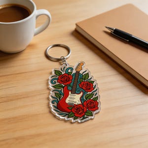 Op de afbeelding: Een heldere acryl sleutelhanger met een rood elektrisch gitaarontwerp, een witte slagplaat en een bruine hals, omringd door rode rozen en groene bladeren. De sleutelhanger is bevestigd aan een zilveren metalen ring.