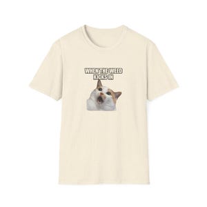 May include: Cream-colored t-shirt featuring a meme of a surprised cat with the text "WHEN THE WEED KICKS IN" in a bold, white font. The cat has orange, white, and brown fur. The shirt has a crew neck and short sleeves.