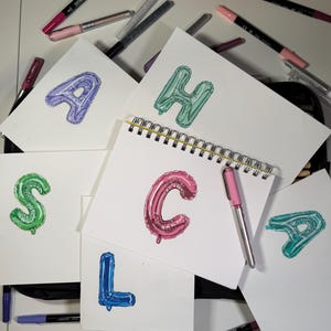 May include: A collection of white cards and a spiral-bound notebook, each featuring a different letter of the alphabet rendered as a colorful balloon. Various acrylic markers in shades of pink, purple, and black are scattered around the cards.