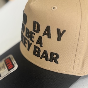 May include: A tan and black baseball cap with the embroidered text "Good Day to Be a Hockey Bar". The hat has a structured crown and a curved brim. The text is in a bold, blocky font.