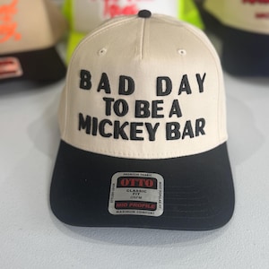 May include: A beige and black baseball cap with the text "BAD DAY TO BE A MICKEY BAR" in black block letters. The cap has a black brim and a small black button on top. The brand tag "OTTO" is visible.