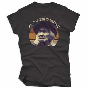 May include: Dark gray t-shirt featuring a graphic of an older person wearing a hat, set against a retro sunset design. The text "HELL IS COMING TO BREAKFAST" is arched above the image. The shirt is a casual wear item.