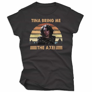 May include: Black t-shirt featuring a graphic of a woman in front of a sunset design. The text "TINA BRING ME THE AXE!" is printed above and below the image. The shirt is made of a soft material.