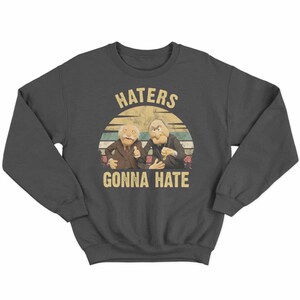 May include: Dark gray sweatshirt featuring a graphic of two puppet characters with the text "HATERS GONNA HATE" in a vintage style. The design includes a retro sunset background.