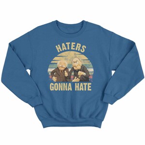 May include: A blue sweatshirt featuring a graphic of two puppet characters with the text "HATERS GONNA HATE" in a vintage style. The design includes a sunset-like background.