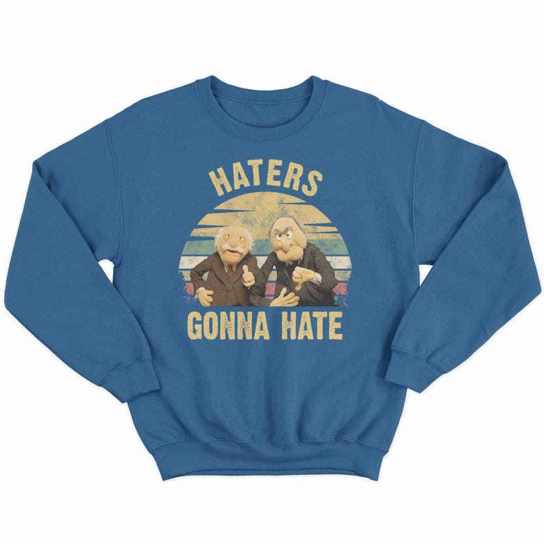 May include: A blue sweatshirt featuring a graphic of two puppet characters with the text "HATERS GONNA HATE" in a vintage style. The design includes a sunset-like background.