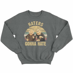 May include: A dark gray sweatshirt featuring a graphic of two Muppet characters with the text "HATERS GONNA HATE" in a vintage style. The design includes a sunset-like background with horizontal stripes.