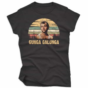 May include: Black t-shirt featuring a retro design with a man's portrait set against a sunset backdrop. The text "GUNGA GALUNGA" is printed below the image in a vintage font. The shirt is made of a soft material.