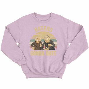 May include: A light purple sweatshirt featuring a graphic of two Muppet characters with the text "HATERS GONNA HATE" in yellow. The graphic includes a sunset design in the background.