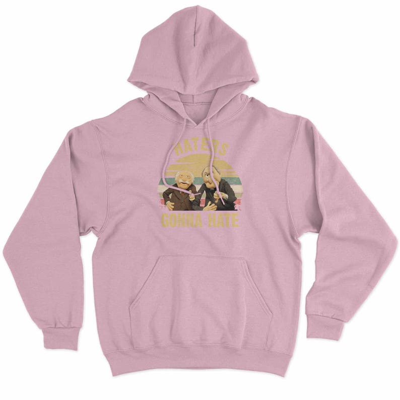 May include: A light pink hoodie with a front pocket and hood. The graphic on the front features two puppet characters with the text "Haters Gonna Hate" in a retro style.