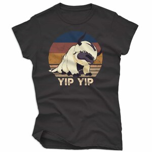 May include: Dark gray t-shirt featuring a cartoon illustration of a flying bison against a retro sunset design. The text "YIP YIP" is printed below the image. The shirt is a crew neck.