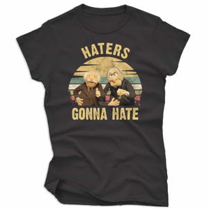 May include: Black t-shirt featuring a graphic of two Muppet characters with the text "HATERS GONNA HATE" in a retro style. The design includes a striped sunset background in shades of yellow, blue, and green.
