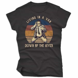 May include: Black t-shirt featuring a retro design with the text "LIVING IN A VAN DOWN BY THE RIVER." The graphic includes a man in a plaid jacket and tie, set against a circular backdrop of blue, orange, and brown.