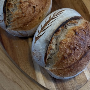 Może przedstawiać: Dwa świeżo upieczone bochenki chleba na zakwasie ze złocistobrązową skórką i dekoracyjnymi nacięciami. Chleb jest prezentowany na drewnianej desce do krojenia, ukazując teksturę i jakość rzemieślniczą. Rustykalna i apetyczna prezentacja.