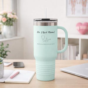 Puede incluir: Un vaso térmico azul claro con asa y pajita. El vaso tiene un borde plateado y una tapa negra. El texto "Dr. [Last Name]" y un gráfico de estetoscopio están impresos en el lateral. El vaso mide aproximadamente 20 cm de alto.