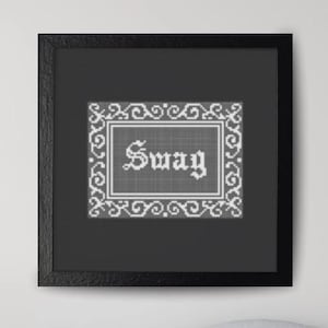 May include: A framed cross-stitch artwork with the word "Swag" in a decorative font. The artwork is in a black frame and hangs on a white wall above a gray sofa. The room has a modern aesthetic with a round coffee table.