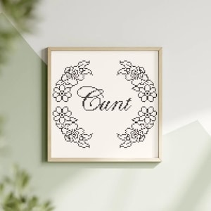 May include: Framed cross-stitch art with the word "Cunt" in the center, surrounded by a floral wreath. The text and floral design are in black on an off-white background, set within a light wood frame.