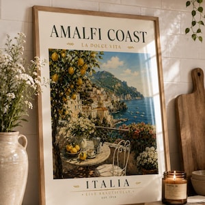 May include: A framed print featuring the Amalfi Coast in Italy. The artwork depicts a scenic view of a coastal town with a lemon tree, a table set for coffee, and the text "AMALFI COAST" and "ITALIA". The print is in a wooden frame.