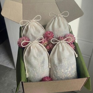 Puede incluir: Una caja de cartón marrón contiene cuatro pequeñas bolsas con cordón de color crema llenas de hierbas secas o té. Flores rosas y papel verde añaden color. La caja la sostiene una persona que lleva una camisa a cuadros rosa y blanca.