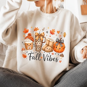 May include: A cream-coloured crewneck sweatshirt featuring a graphic print of a coffee cup with a beanie, a leopard print tumbler with a bow, tan boots, and a pumpkin with a plaid bow. The text Fall Vibes is printed in black cursive script.