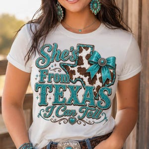 Puede incluir: Camiseta color crema con el texto "She's From TEXAS I Can Tell" en letras turquesas brillantes. El dise&ntilde;o incluye un gr&aacute;fico en forma de Texas con un estampado de piel de vaca y un lazo turquesa. La camiseta tiene mangas cortas.