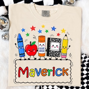 May include: A beige t-shirt featuring a graphic of a blue crayon, a red apple, an orange glue stick, a black and white composition notebook, and a yellow pencil, all with smiling faces. Below them is a scalloped box with the name Maverick in colours.