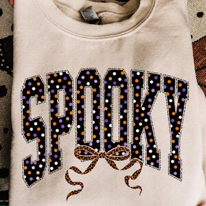 May include: A beige crewneck sweatshirt featuring the word SPOOKY in large block letters. The letters have a black background with orange, purple, and white polka dots. A brown leopard print bow graphic is positioned below the text.