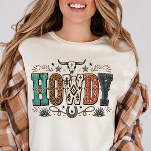 May include: A cream-colored t-shirt featuring the word HOWDY in large, patterned letters. The design includes a steer skull, stars, a horseshoe with rope, and agave plants in shades of turquoise, brown, cream, red, and dark blue.