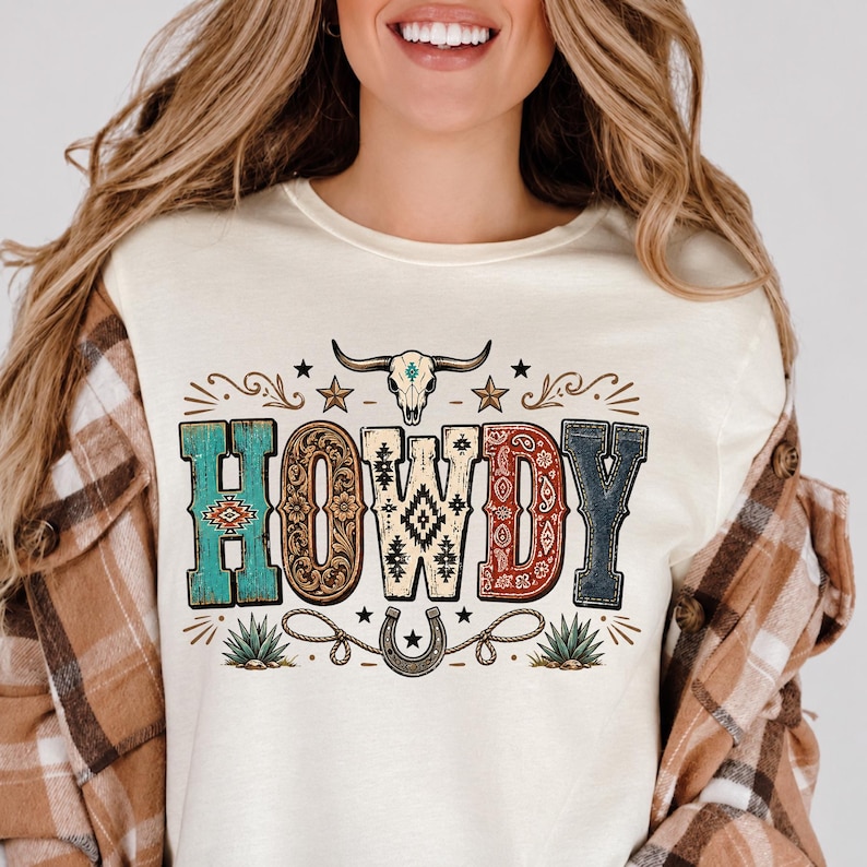 May include: A cream-colored t-shirt featuring the word HOWDY in large, patterned letters. The design includes a steer skull, stars, a horseshoe with rope, and agave plants in shades of turquoise, brown, cream, red, and dark blue.