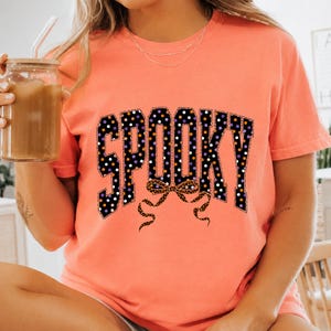 May include: A coral-coloured short-sleeve t-shirt featuring the word SPOOKY in large block letters. The letters are black with a white, orange, and purple polka dot pattern. Below the text is a leopard print bow graphic with long flowing ribbons.