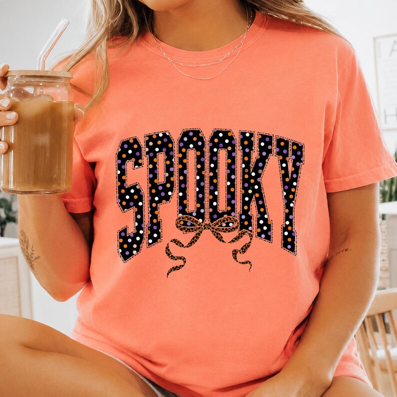 May include: A coral-coloured short-sleeve t-shirt featuring the word SPOOKY in large block letters. The letters are black with a white, orange, and purple polka dot pattern. Below the text is a leopard print bow graphic with long flowing ribbons.