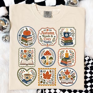 以下が含まれることがあります： クリーム色のTシャツで、9つの装飾的なイラストがグリッド状に配置されています。中央のグラフィックには「Autumn Reads & Cozy Nights」という文字があり、その周りにカボチャ、本、キノコ、キャンドル、コーヒーマグ、秋の葉のイラストが描かれています。