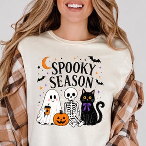 May include: A cream-colored t-shirt featuring a graphic with the text SPOOKY SEASON in black. The design includes a white ghost holding an orange drink, a skeleton, a jack-o'-lantern, a black cat with a purple bow, bats, and orange stars.