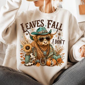 May include: A cream-colored crewneck sweatshirt featuring a graphic of a Highland cow wearing a teal cowboy hat and sunglasses. The text reads LEAVES FALL I DON'T. The design includes a sunflower, a cowboy boot, a horseshoe, and pumpkins.