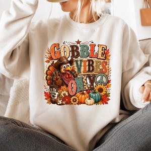 May include: A cream-colored crewneck sweatshirt featuring a graphic print of a turkey wearing a cowboy hat and bandana, making a peace sign. The text reads GOBBLE VIBES ONLY, surrounded by sunflowers, pumpkins, and autumn leaves in warm tones.