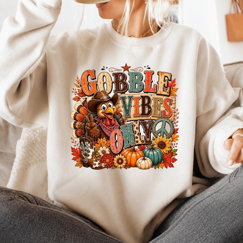 May include: A cream-colored crewneck sweatshirt featuring a graphic print of a turkey wearing a cowboy hat and bandana, making a peace sign. The text reads GOBBLE VIBES ONLY, surrounded by sunflowers, pumpkins, and autumn leaves in warm tones.