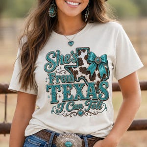 Puede incluir: Camiseta color crema con el texto "She's From Texas I Can Tell" en letras brillantes turquesas. El dise&ntilde;o incluye un contorno en forma de Texas con un estampado de piel de vaca y un lazo turquesa.