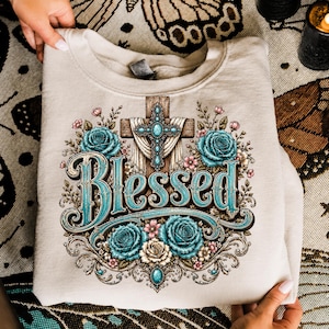 May include: A beige crewneck sweatshirt featuring a graphic print of a wooden cross adorned with turquoise stones and draped fabric, surrounded by teal and cream roses. The word Blessed is printed in large, ornate teal and brown lettering below.