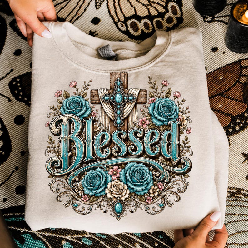 May include: A beige crewneck sweatshirt featuring a graphic print of a wooden cross adorned with turquoise stones and draped fabric, surrounded by teal and cream roses. The word Blessed is printed in large, ornate teal and brown lettering below.