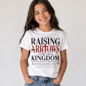 Peut inclure: Un t-shirt blanc avec l'inscription "RAISING ARROWS FOR THE KINGDOM" en noir et rouge. Le t-shirt comprend &eacute;galement une citation du Psaume 127:4-5. Port&eacute; par une personne aux cheveux bruns et un jean bleu.
