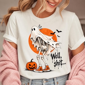 May include: A white t-shirt featuring a graphic of a skeleton wearing a ghost sheet and orange sneakers, standing next to a jack-o'-lantern in front of a large orange moon. The design includes black bats and the text "Well, shit..." in a dripping font.