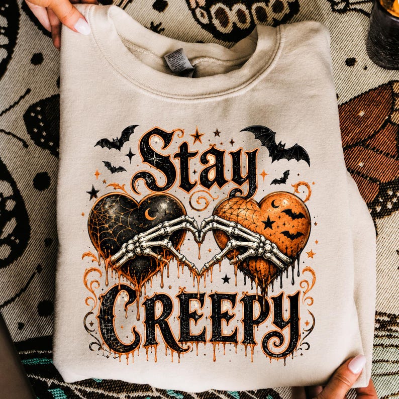 May include: A beige crewneck sweatshirt featuring a graphic print with the text Stay CREEPY. The design includes two hearts, one black with a spiderweb and one orange with bats, joined by two skeleton hands forming a heart shape with flying bats.