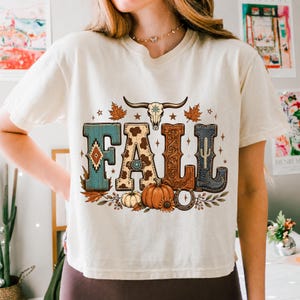 May include: A cream-colored short-sleeve t-shirt featuring a graphic print of the word FALL. The letters are decorated with turquoise patterns, cow print, floral carvings, and a cowboy boot. A bull skull, pumpkins, and leaves surround the text.