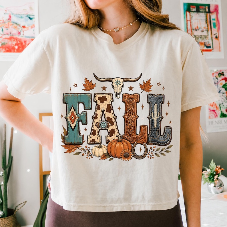 May include: A cream-colored short-sleeve t-shirt featuring a graphic print of the word FALL. The letters are decorated with turquoise patterns, cow print, floral carvings, and a cowboy boot. A bull skull, pumpkins, and leaves surround the text.