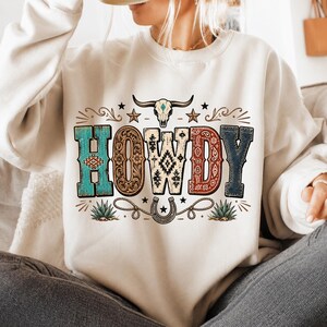 May include: A cream-colored crewneck sweatshirt featuring the word HOWDY in large, multi-patterned letters. Above the text is a steer skull, and below is a horseshoe and rope graphic. The design includes turquoise, brown, and red accents.
