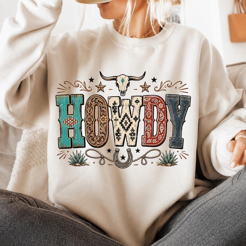 May include: A cream-colored crewneck sweatshirt featuring the word HOWDY in large, multi-patterned letters. Above the text is a steer skull, and below is a horseshoe and rope graphic. The design includes turquoise, brown, and red accents.